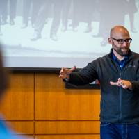 man gesturing as he talks at the front of a room, black and white photo projected behind him